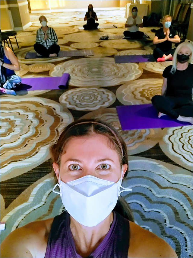 Selfie of Adriane Dellorco wearing a face mask, with people on yoga mats visible in the background.