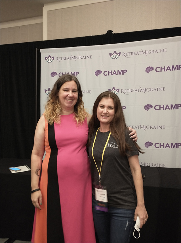  Two women with their arms around each other's shoulders in front of a black curtain and CHAMP RetreatMigraine photo backdrop.