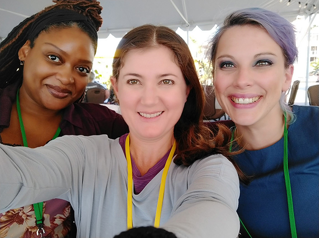 Adriane Dellorco between two smiling friends sitting under a white event tent outdoors.