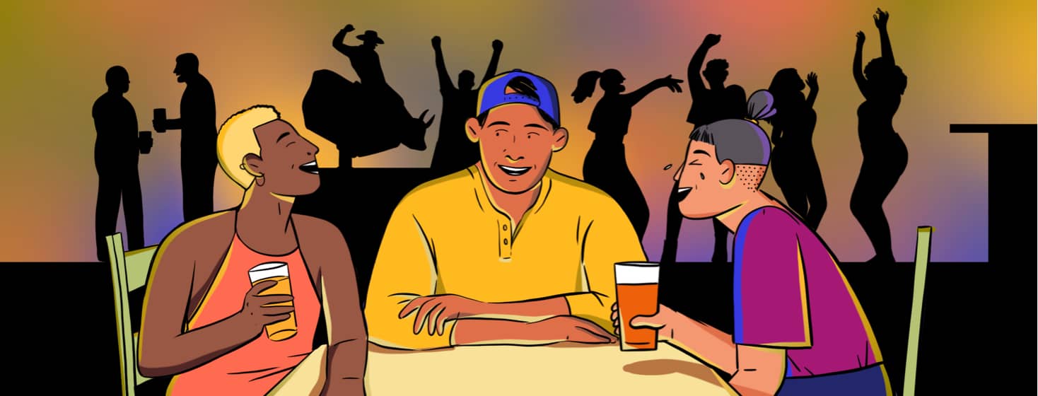 A man tells a funny story to people sitting at a table with him while people dance, drink, and ride a mechanical bull in the background.